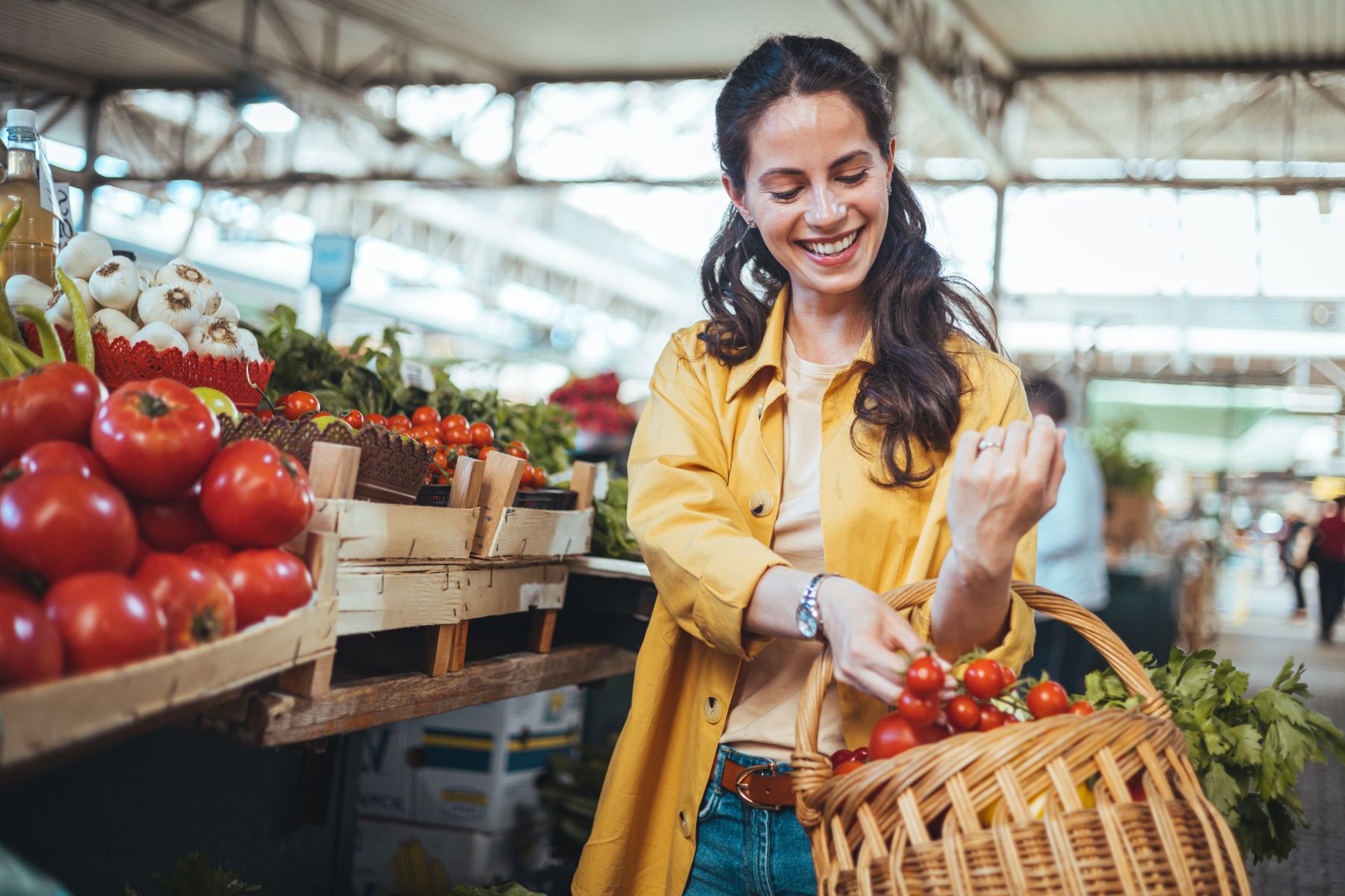 Woman in a yellow coat shops for tomatoes at a farmer's market, smiling as she fills a wicker basket with red cherry tomatoes.